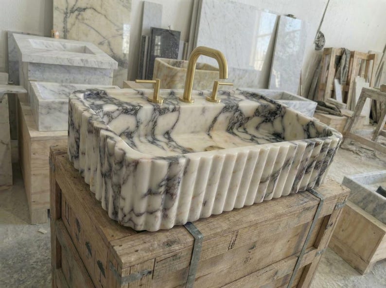 Puede incluir: Un lavabo rectangular de m&aacute;rmol con un patr&oacute;n veteado blanco y gris. El lavabo tiene un exterior acanalado y grifos dorados. El lavabo se exhibe sobre una caja de madera en un taller.