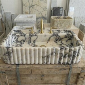 Puede incluir: Un lavabo rectangular de m&aacute;rmol con exterior estriado y grifer&iacute;a dorada. El lavabo presenta una base blanca con vetas gris oscuro. Se exhibe sobre una caja de madera, con otros lavabos de m&aacute;rmol al fondo.