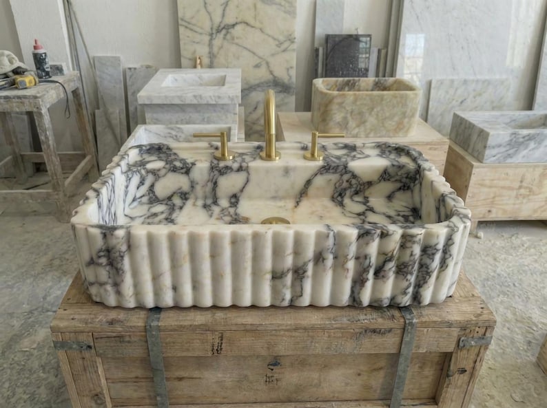 Puede incluir: Un lavabo rectangular de m&aacute;rmol con exterior estriado y grifer&iacute;a dorada. El lavabo presenta una base blanca con vetas gris oscuro. Se exhibe sobre una caja de madera, con otros lavabos de m&aacute;rmol al fondo.