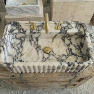 Puede incluir: Un lavabo rectangular de m&aacute;rmol con borde festoneado, con un patr&oacute;n veteado blanco y gris. El lavabo tiene grifos y herrajes de desag&uuml;e dorados. Est&aacute; colocado sobre una caja de madera.