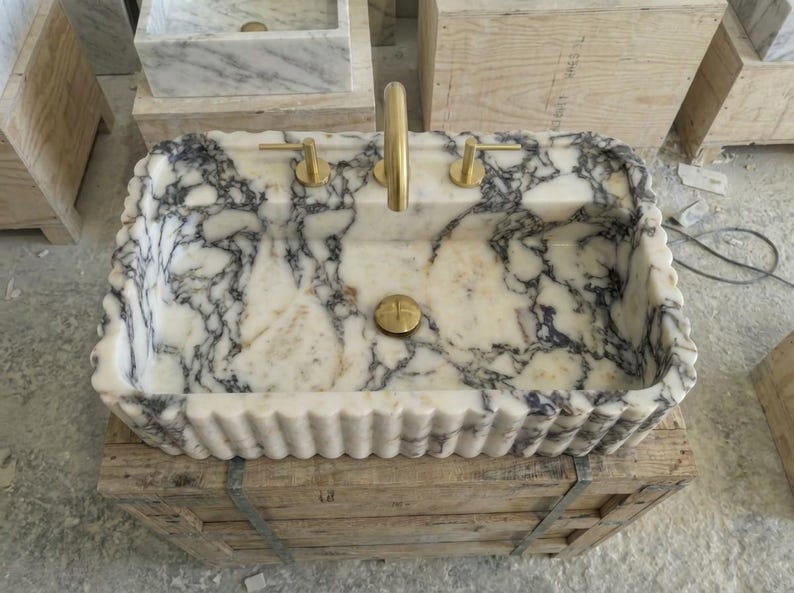 Puede incluir: Un lavabo rectangular de m&aacute;rmol con borde festoneado, con un patr&oacute;n veteado blanco y gris. El lavabo tiene grifos y herrajes de desag&uuml;e dorados. Est&aacute; colocado sobre una caja de madera.