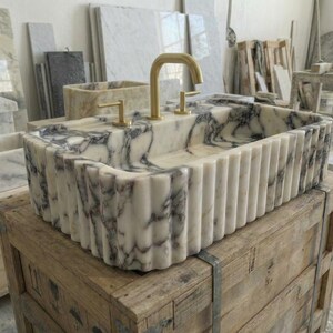Puede incluir: Un lavabo rectangular de m&aacute;rmol con exterior estriado y grifos dorados. El lavabo presenta un patr&oacute;n veteado blanco y gris. Se exhibe sobre una caja de madera en un taller.
