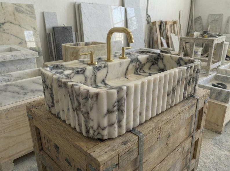 Puede incluir: Un lavabo rectangular de m&aacute;rmol con exterior estriado y grifos dorados. El lavabo presenta un patr&oacute;n veteado blanco y gris. Se exhibe sobre una caja de madera en un taller.