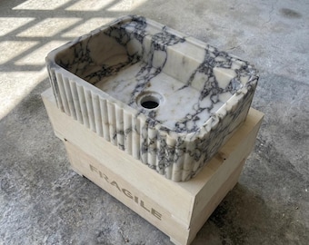 May include: A rectangular marble sink with a white and gray veined pattern. The sink has a fluted exterior and a circular drain. It rests on a wooden crate with the word "FRAGILE" printed on the side.