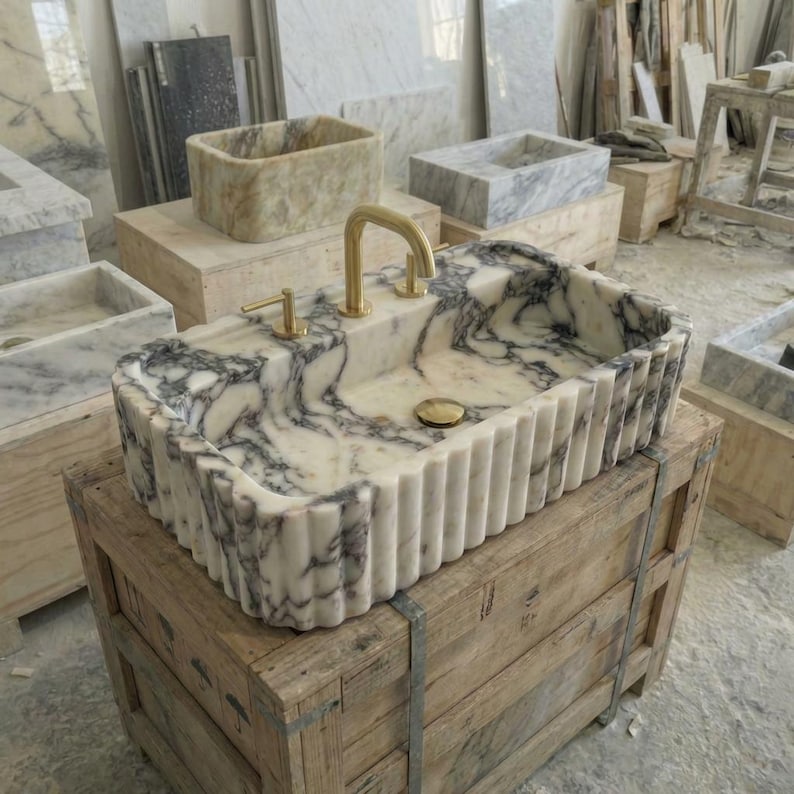Puede incluir: Un lavabo rectangular de m&aacute;rmol con exterior estriado y grifo dorado. El lavabo presenta un patr&oacute;n veteado blanco y gris. Otros lavabos de m&aacute;rmol son visibles en el fondo, apoyados sobre cajas de madera.