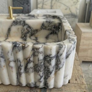 Puede incluir: Un lavabo rectangular de m&aacute;rmol con borde acanalado. El lavabo tiene una base blanca con vetas grises y grifos dorados. El lavabo es de piedra natural y es un accesorio de ba&ntilde;o.
