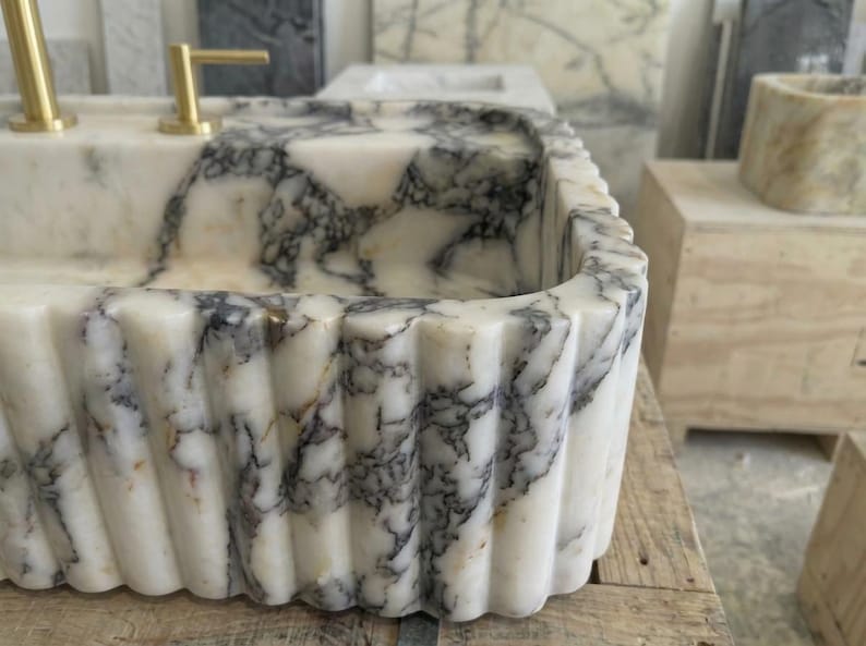 Puede incluir: Un lavabo rectangular de m&aacute;rmol con borde acanalado. El lavabo tiene una base blanca con vetas grises y grifos dorados. El lavabo es de piedra natural y es un accesorio de ba&ntilde;o.