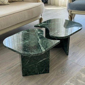May include: Two green marble side tables with distinctive, curved designs. The tables are dark green with white veining, supported by rectangular bases. They are positioned on a light wood floor. The tables are a stylish addition.