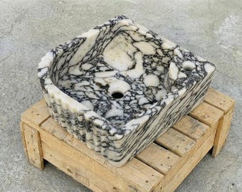 Può includere: Un lavabo quadrato in marmo caratterizzato da un sorprendente motivo venato bianco e nero. Le pareti esterne del bacino presentano una texture scanalata verticale, mentre l'interno include un foro di scarico centrale e un piccolo foro circolare.
