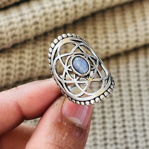 Könnte beinhalten: Ein silberfarbener Ring mit einem ovalen, hellblauen Cabochon-Stein in der Mitte eines komplexen, durchbrochenen Metallrahmens mit Perlenrand. Das Design besteht aus überlappenden kreisförmigen Mustern, die den zentralen Stein umgeben.