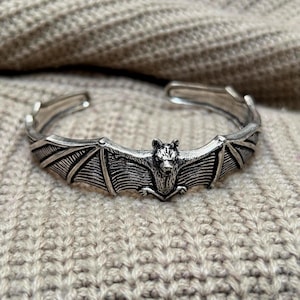 Può includere: Bracciale a polsino argentato con un design dettagliato a pipistrello. Il pipistrello è al centro, con ali spiegate e dettagli intricati. Il bracciale ha un design aperto per una facile indossabilità.