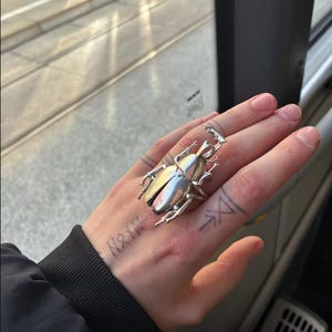 May include: A silver beetle ring with detailed wings and body. The ring is worn on a hand with tattoos, including text and geometric designs. The ring's design is intricate and eye-catching.
