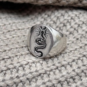 Puede incluir: Anillo de sello plateado con un diseño detallado de serpiente negra dentro de una forma ovalada. La serpiente está colocada debajo de un símbolo de luna creciente y sol. El anillo está sobre un fondo gris claro texturizado.