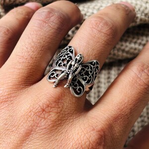 May include: A silver-colored butterfly ring with intricate filigree wings and a central figure. The ring is worn on a finger, showcasing the detailed design and metallic sheen. The butterfly's wings are open, creating a delicate and ornamental piece.