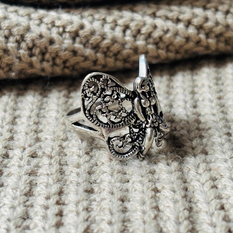 May include: A silver-colored ring with a detailed fairy design. The ring features a filigree wing and a sculpted fairy figure. The ring is set against a textured, light-colored background, showcasing the intricate details of the jewelry.