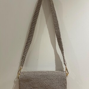 May include: A beige crochet shoulder bag with a rectangular body and a long strap. The bag has a gold-coloured clasp and hardware. The bag is hanging against a plain white wall.