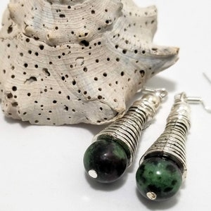 Peut inclure: Une paire de boucles d'oreilles pendantes en argent avec des perles de pierres précieuses vertes. Les boucles d'oreilles ont un design tissé et sont accentuées par une perle d'argent en haut.