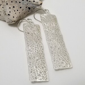 May include: A pair of silver dangle earrings with a delicate, textured design. The earrings are rectangular in shape and have a slightly curved, organic look.