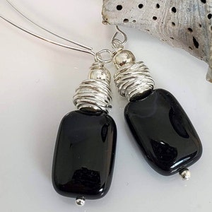 May include: A pair of black rectangular earrings with silver wire detailing. The earrings have a polished, glossy finish and are suspended from thin silver wires. The design includes silver beads and coiled wire accents, creating a modern, minimalist aesthetic.
