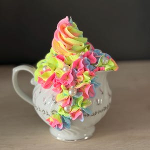 Puede incluir: Una cremera de porcelana blanca con detalles florales, adornada con glaseado vibrante y multicolor en tonos rosa, amarillo y verde. El glaseado está decorado con cuentas perladas, creando una pieza caprichosa y decorativa.