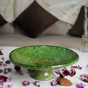 Op de afbeelding: Een groene keramische voetbakschaal met een getextureerd oppervlak en een decoratieve rand. De schaal staat op een wit oppervlak, omringd door gedroogde bloemen en bloemblaadjes. De diameter van de schaal is ongeveer 30 cm.