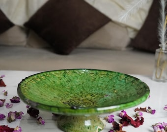 May include: A green ceramic pedestal bowl with a textured surface and a decorative rim. The bowl is placed on a white surface, surrounded by dried flowers and petals. The bowl's diameter is approximately 12 inches.
