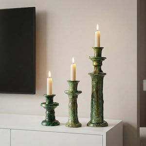 Könnte beinhalten: Drei grüne Kerzenhalter aus Keramik in verschiedenen Höhen, die jeweils eine brennende, cremefarbene Kerze halten. Die Halter haben ein strukturiertes, gealtertes Aussehen mit glänzendem Finish. Der höchste Halter ist etwa 30 cm hoch.