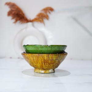 Può includere: Due ciotole in ceramica impilate. La ciotola superiore è di un verde vibrante, mentre la ciotola inferiore è di un giallo-marrone strutturato. Lo sfondo sfocato presenta un vaso bianco e piante secche, creando un'estetica rustica.