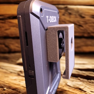 May include: A close-up of a grey T-DECK device with a light brown bracket attached. The device has a dark grey body with the text "T-DECK" printed on it. The bracket is attached to the side of the device and has a rectangular shape.