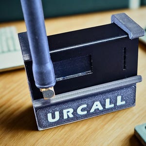 Könnte beinhalten: Ein schwarzes, rechteckiges elektronisches Gerät mit einer vertikalen Antenne an der linken Seite. Die Vorderseite verfügt über ein strukturiertes Panel mit dem Wort URCALL in weißen Großbuchstaben. Das Gerät hat ein mattes, kompaktes Design.