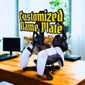 以下が含まれることがあります： 黄色い文字でCustomized Name Plateと書かれた看板が上部にある、黒いゲーミングアクセサリースタンド。垂直の支柱には、2つの黒いヘッドセットと2つの白いゲームコントローラーを掛けるためのフックが付いています。