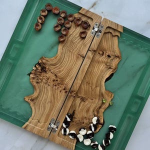 May include: Backgammon board with a green resin border and a wooden playing surface. The board is open, revealing the wooden interior with brown and white game pieces. The board is square and has metal hinges.