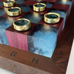 Może przedstawiać: Ręcznie wykonana szachownica z ramą z ciemnego drewna i naprzemiennymi kwadratami z ciemnoczerwonego drewna i niebieskiej żywicy. Złote pionki z ciemnozielonymi środkami umieszczone są na planszy. Litery "G" i "H" są wyryte w ramie.