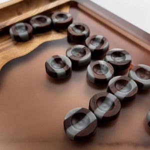 May include: A collection of backgammon game pieces. The pieces are dark brown and gray, with a circular shape and a striped design. They are arranged on a wooden backgammon board.