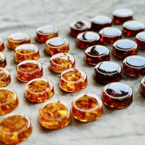 Puede incluir: Colección de pequeños objetos redondos de vidrio color ámbar con interior texturizado. Algunos tienen una parte superior oscura y reflectante, otros son más claros. Los objetos están dispersos sobre una superficie gris.