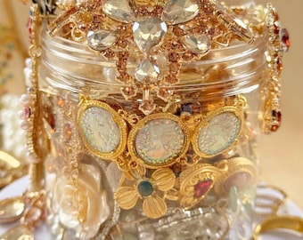 May include: A clear glass jar overflowing with an assortment of gold-toned jewelry. The jar is topped with a decorative lid adorned with large, clear gemstones and pearls. Various rings, necklaces, and earrings are visible, creating a luxurious and ornate display.