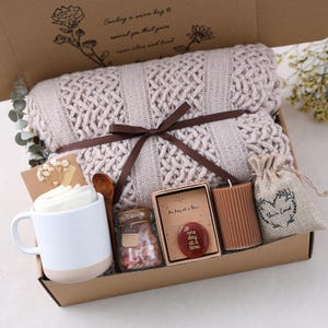 May include: A gift box with a beige knitted throw blanket tied with a brown ribbon. It includes a white mug, a wooden spoon, a candle, a small bag, and a jar of bath salts. The box has text that reads "Sending a warm hug to remind you that you're never alone and loved."