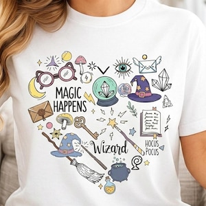 May include: White t-shirt featuring a heart-shaped design with whimsical illustrations. The design includes glasses, a crystal ball, a witch's hat, and the words "MAGIC HAPPENS" and "WIZARD". Additional text reads "HOCUS POCUS".