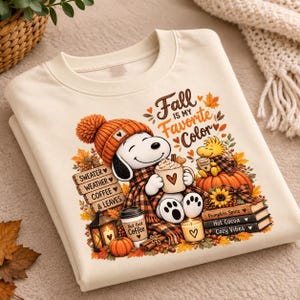 Puede incluir: Una sudadera color crema con un gráfico de Snoopy con un gorro de punto naranja y una bufanda a cuadros, sosteniendo una taza. El texto dice Fall is my Favorite Color, Sweater Weather, Coffee & Leaves, But first Coffee, Pumpkin Spice, Hot Cocoa, Cozy Vibes.