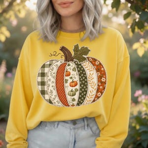 Könnte beinhalten: Ein senfgelbes Langarm-Sweatshirt mit einer großen zentralen Grafik eines Kürbisses, der aus verschiedenen vertikalen Paneelen besteht. Die Paneele zeigen unterschiedliche Muster wie grünes Vichy-Karo, Blumenprints und Herbstblätter.