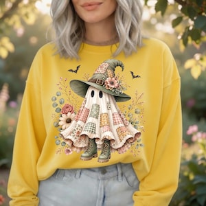 May include: A mustard yellow long-sleeve sweatshirt featuring a central graphic of a ghost wearing a patchwork witch hat and boots. The ghost is surrounded by floral elements and small bats, with a patchwork pattern covering its body.