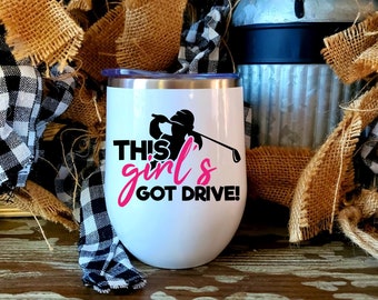 Op de afbeelding: Een witte roestvrijstalen tumbler met een zwart silhouet van een persoon die golf speelt en de tekst "This girl's got drive!" in roze en zwarte letters.