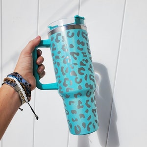 May include: A turquoise stainless steel travel mug with a leopard print design and a handle. The mug has a lid with a flip top.