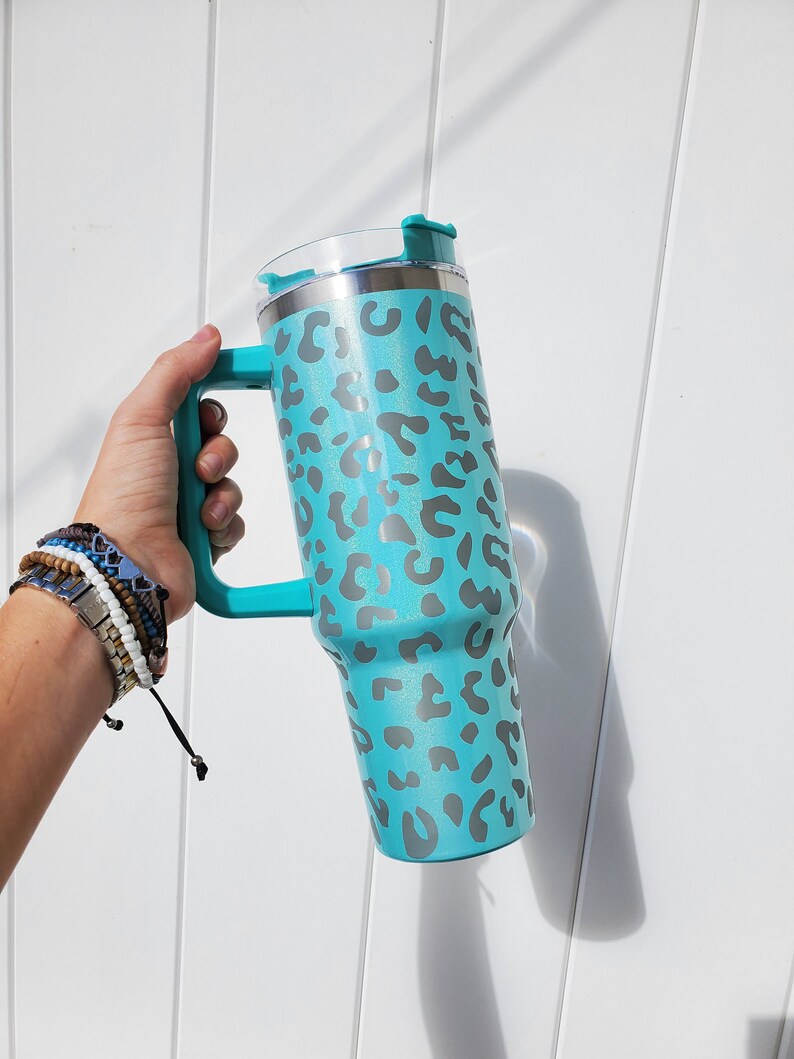 May include: A turquoise stainless steel travel mug with a leopard print design and a handle. The mug has a lid with a flip top.