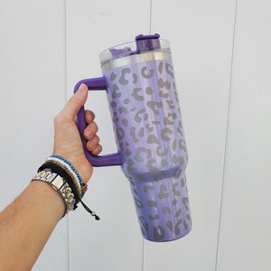 May include: A purple stainless steel tumbler with a leopard print design and a lid. The tumbler has a handle and is being held by a hand.