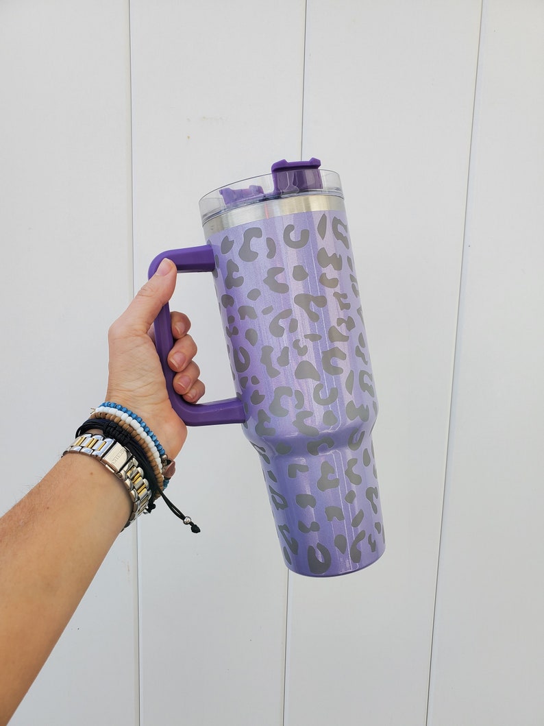 May include: A purple stainless steel tumbler with a leopard print design and a lid. The tumbler has a handle and is being held by a hand.
