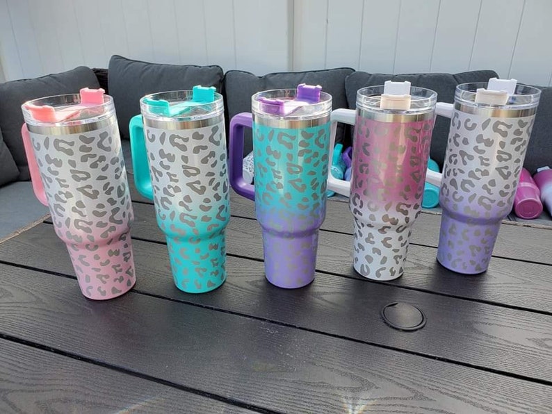 May include: Five stainless steel travel mugs with a leopard print design. The mugs are in different colors: pink, teal, purple, white, and lavender. Each mug has a lid with a handle.