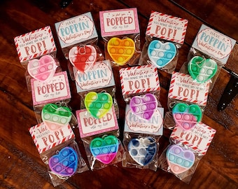 Op de afbeelding: Een verzameling hartvormige Pop It fidget-speeltjes in verschillende kleuren, verpakt met Valentijnsdagkaarten. De kaarten bevatten zinnen als "You Make My Heart POP!" en "Have a Poppin' Valentine's Day!"