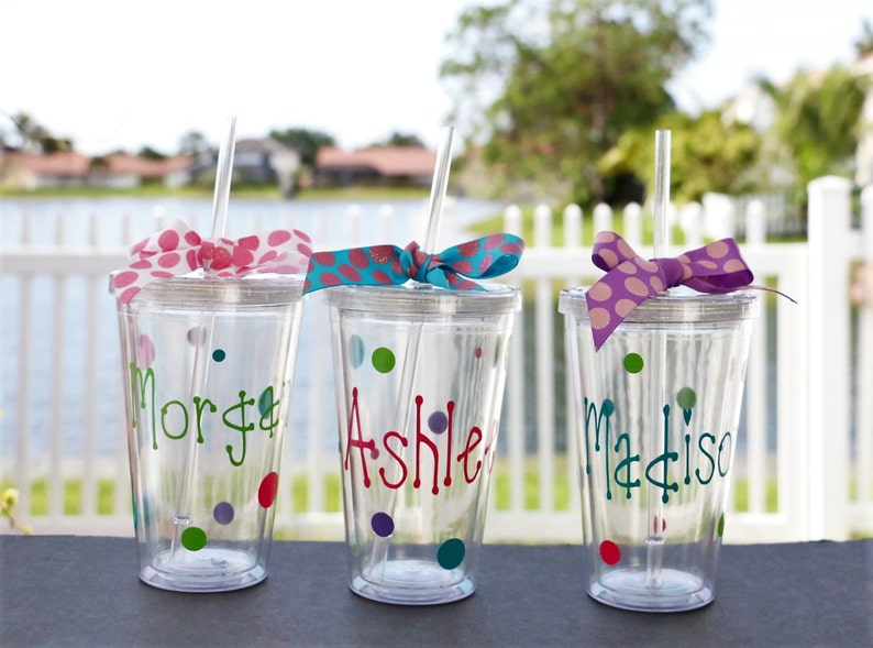 Op de afbeelding: Drie doorzichtige plastic bekers met rietjes en gepersonaliseerde namen, "Morgan", "Ashle" en "Madison", versierd met kleurrijke stippen en strikken.