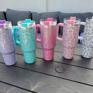 May include: Five stainless steel travel mugs with a leopard print design. The mugs are in different colors: purple, teal, pink, light pink, and white. Each mug has a handle and a lid.